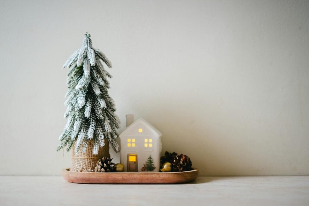 A minimalist Christmas scene with a small snowy tree, lit house, and pinecones on a tray indoors.